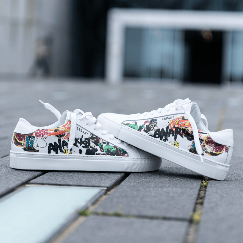 Et par hvide sneakers med farverigt graffiti-design og teksten "Anarkist" på siden, placeret på en udendørs overflade.