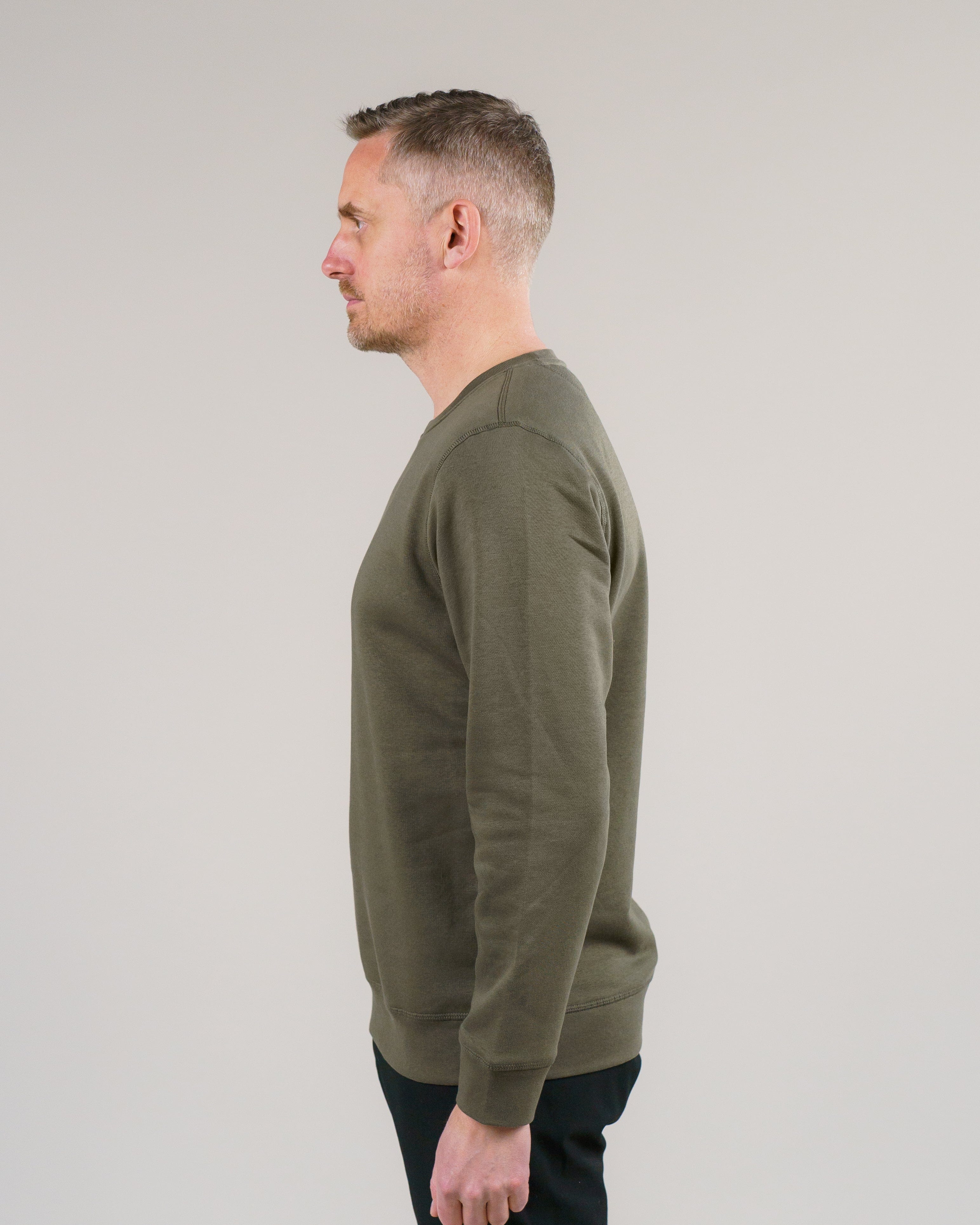 En mand iført en olivengrøn sweatshirt står i profil mod en neutral baggrund.